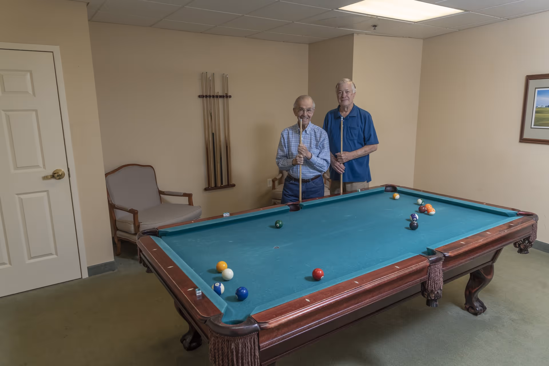 Two elderly men standing behind a pool table in a room with beige walls. They are holding pool cues and smiling. There is a chair and a rack with pool cues on the wall behind them, and a framed picture on the right wall.