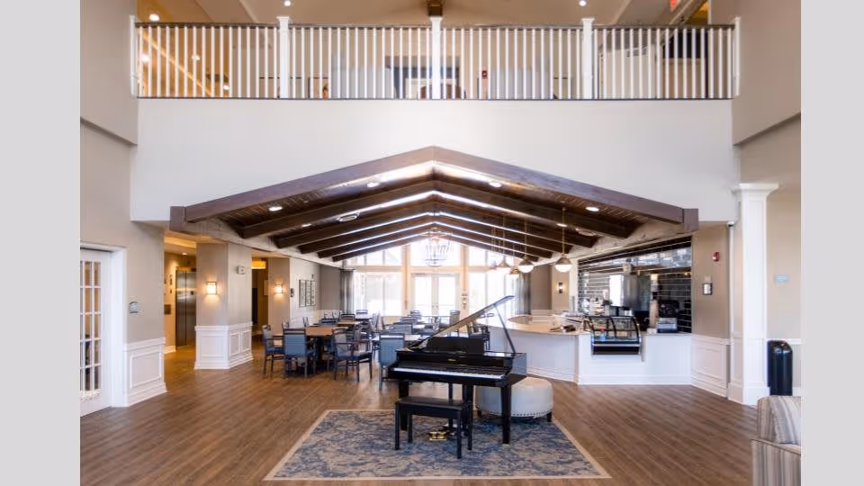 Spacious senior community common area with a grand piano in the foreground, dining tables, and a vaulted beamed ceiling.