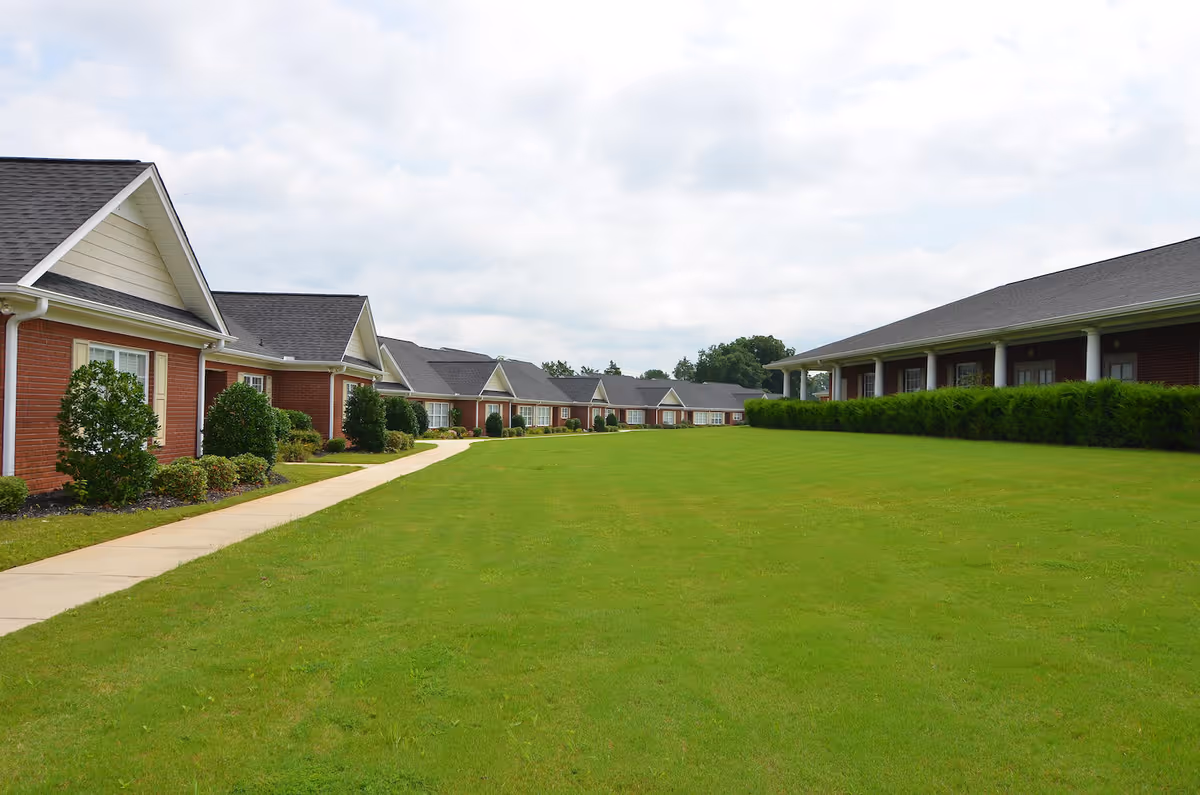 A wide view of a senior living facility with a large green lawn in the center, bordered by single-story brick buildings with white trim and dark roofs on both sides. A concrete walkway runs along the left side of the lawn, and there are bushes and small trees planted near the buildings. The sky is partly cloudy.