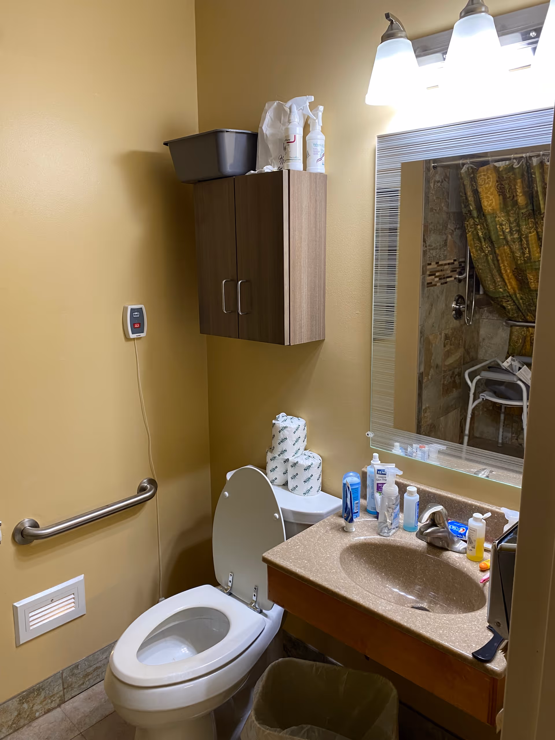 A bathroom with a toilet, a sink with various toiletries on the countertop, a wall-mounted cabinet above the toilet, and a mirror reflecting a shower area with a patterned shower curtain and a shower chair. The walls are painted beige, and there is a grab bar next to the toilet.