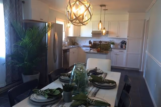 A modern kitchen and dining area featuring white cabinetry, stainless steel appliances including a refrigerator and stove, and a white dining table set with plates, green napkins, and decorative plants. The room is illuminated by a spherical chandelier and pendant lights, with a large window covered by a curtain on the left side.