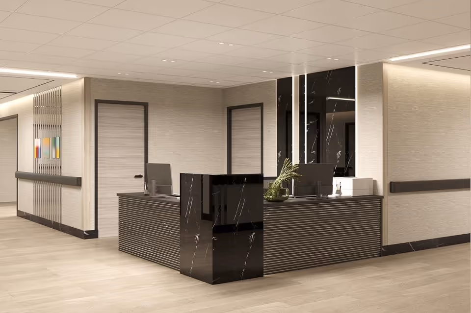 A modern nursing home reception desk area with a black marble counter and two computer monitors. The walls are beige with a textured finish, and there are two closed doors behind the desk. The floor is light wood, and the ceiling has recessed lighting. A small plant and some office supplies are on the desk.
