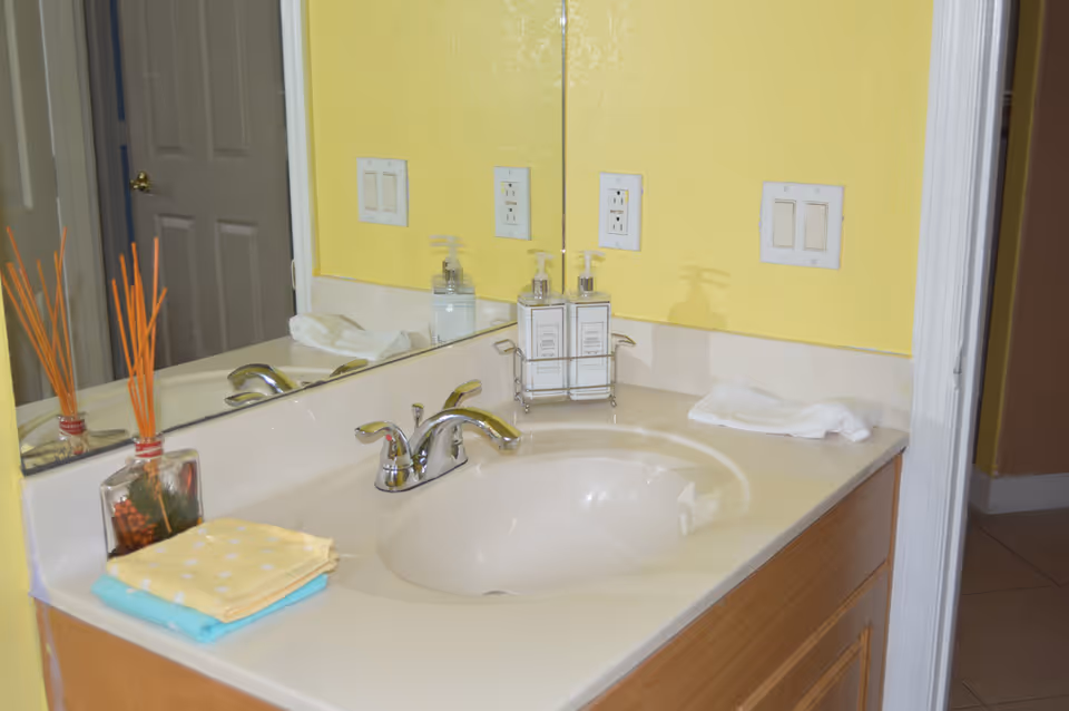 Bathroom vanity with an oval sink, chrome faucet, mirror, soap dispensers, folded towels and a yellow wall.