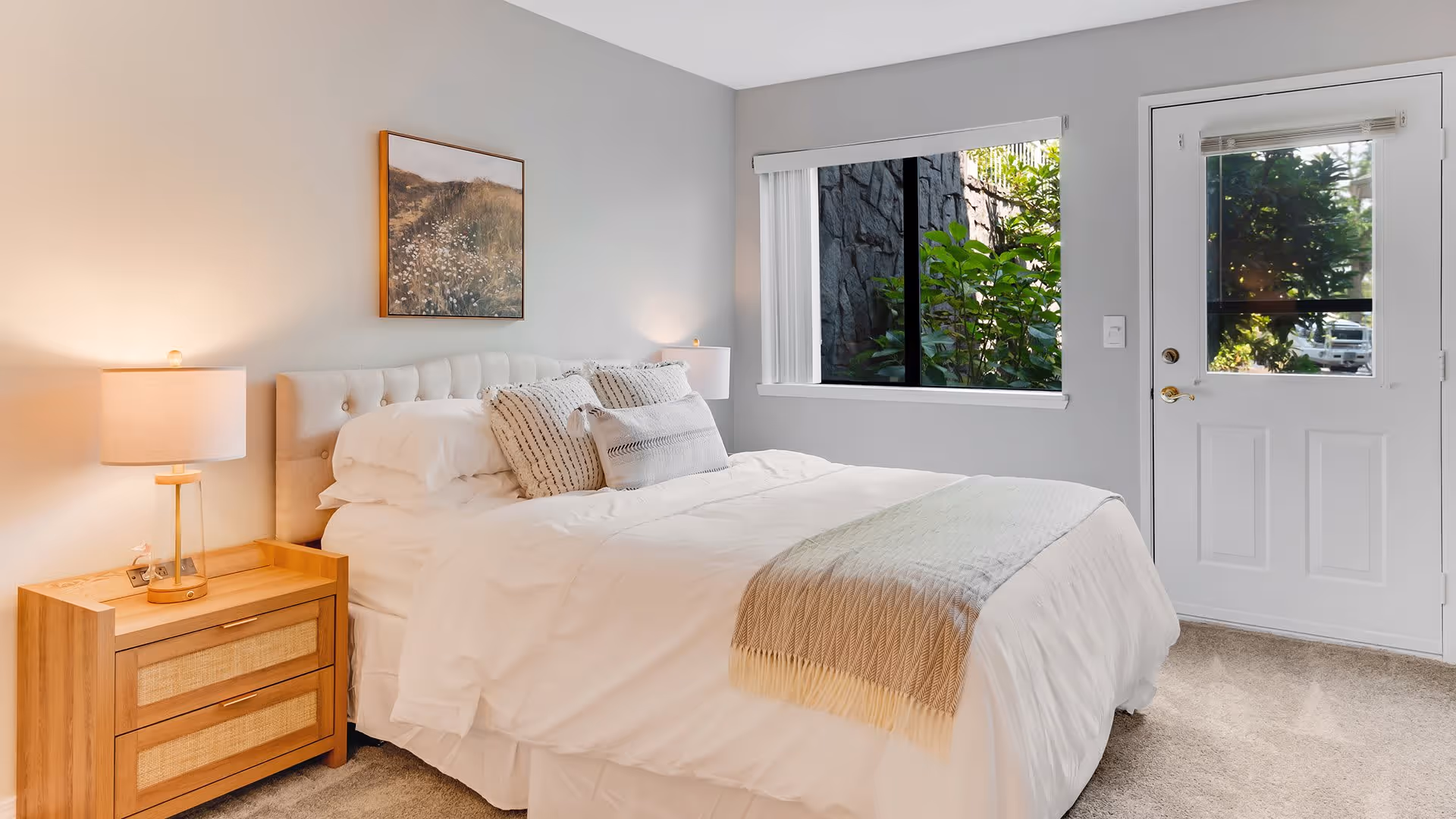 A bright and cozy bedroom with a neatly made bed featuring white linens and decorative pillows. There is a wooden nightstand with a lamp on the left side of the bed. A framed picture hangs on the light gray wall above the bed. A window and a white door with glass panels are on the right side, showing greenery outside.