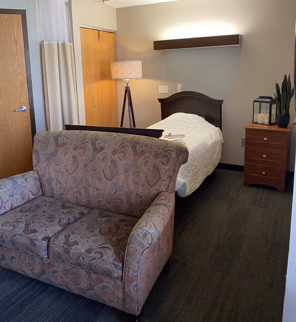 A small furnished bedroom in a senior living facility with a single bed, bedside table and lamp, and a patterned loveseat in the foreground.