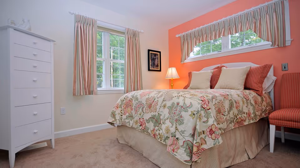 Cozy bedroom with a floral bedspread and striped pillows against a coral accent wall, a white tall dresser, bedside lamp, and windows with striped curtains.