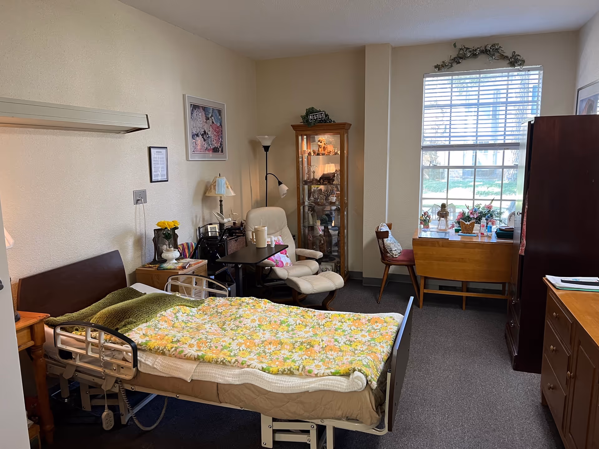 Furnished senior living bedroom containing an adjustable bed with a floral blanket, a recliner and side tables, a glass display cabinet, and a window with a small table.