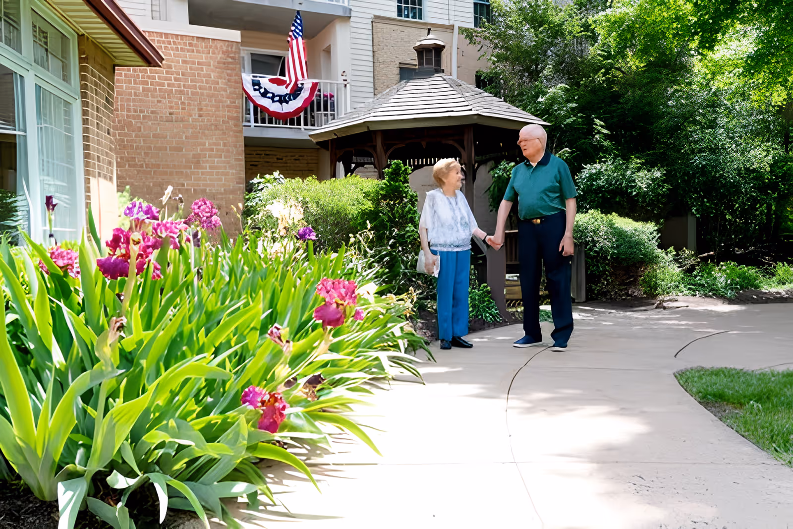 An elderly couple holding hands and smiling at each other while standing on a curved pathway in a garden area outside a building. The garden has blooming purple flowers and green bushes, with a wooden gazebo in the background. An American flag and patriotic bunting are displayed on the building.