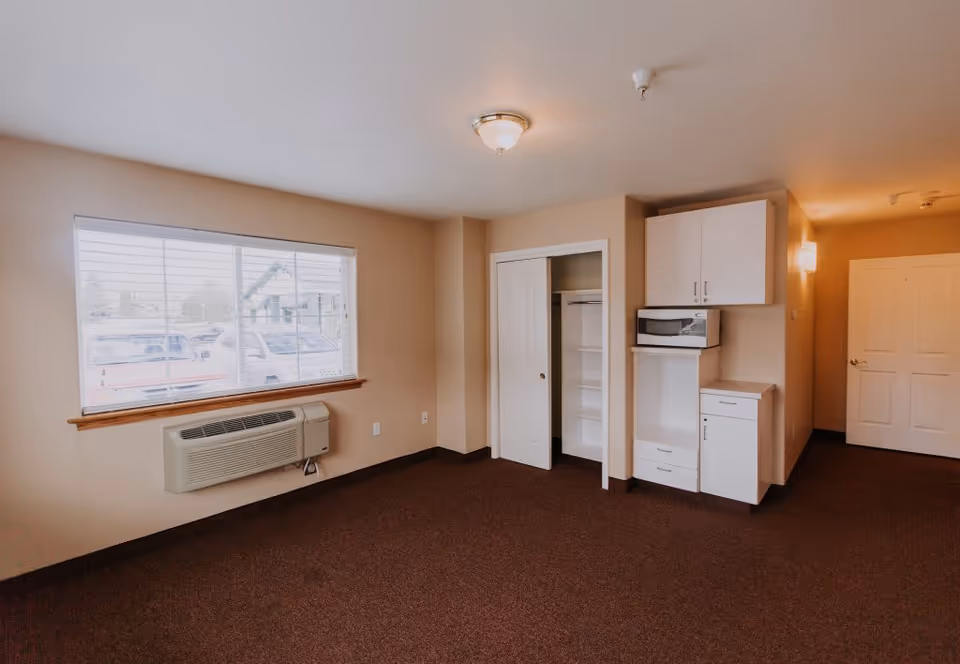Empty room in an assisted living facility with beige walls, a large window with blinds showing parked cars outside, a wall-mounted air conditioning unit below the window, an open closet with sliding doors, and a small kitchenette area with white cabinets and a microwave.
