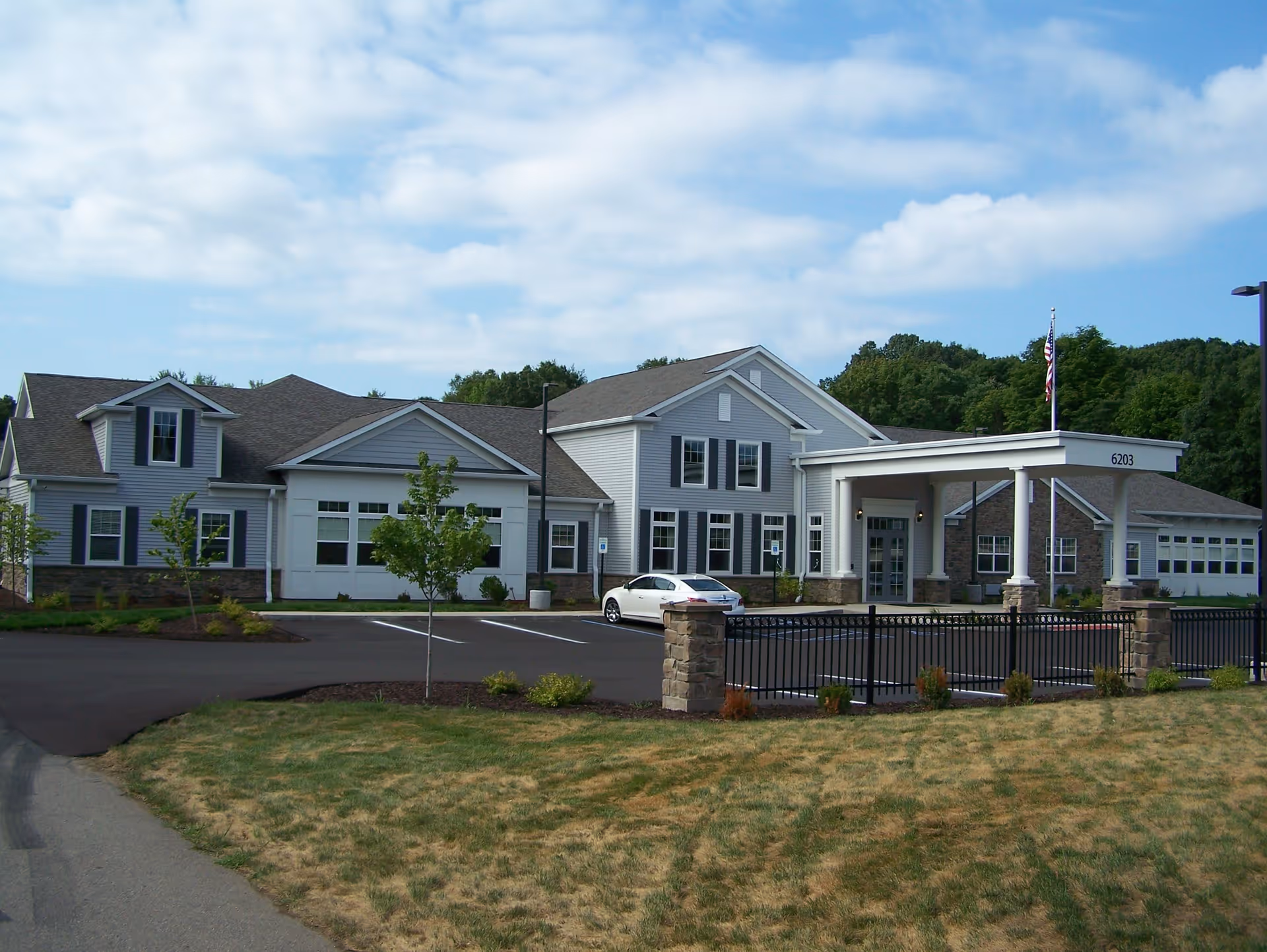 Exterior view of a single-story senior living facility building with gray siding, white trim, and a covered entrance. There is a parking lot with a white car, a small fenced area with stone pillars, and some young trees and landscaping in the foreground. The sky is partly cloudy.