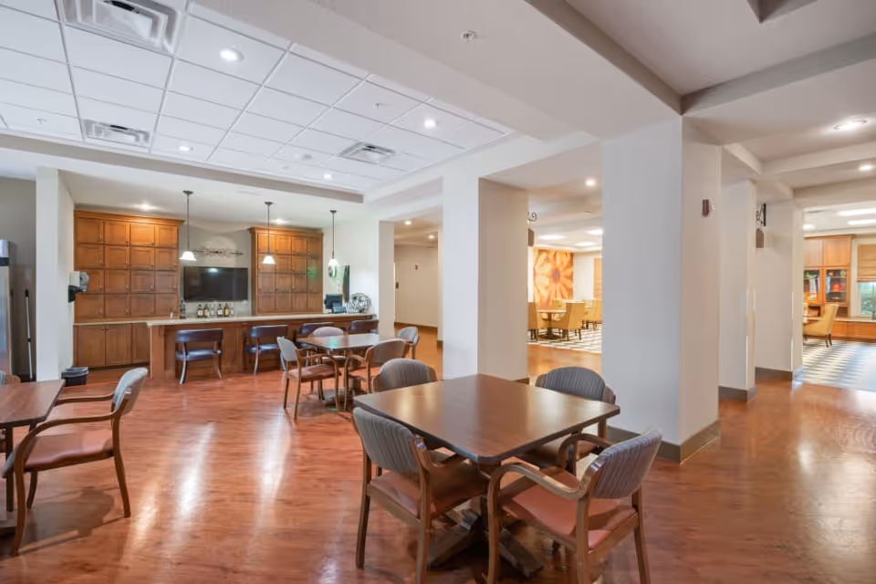 Interior view of a senior living facility common area with wooden tables and chairs arranged on a polished wooden floor. In the background, there is a bar area with wooden cabinets, a countertop, and three pendant lights hanging from the ceiling. The space is well-lit with recessed lighting and has a clean, modern design.