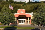 Front exterior view of a community center building with a red and beige facade, large glass entrance doors, an American flag on a flagpole to the left, and trees lining the front.