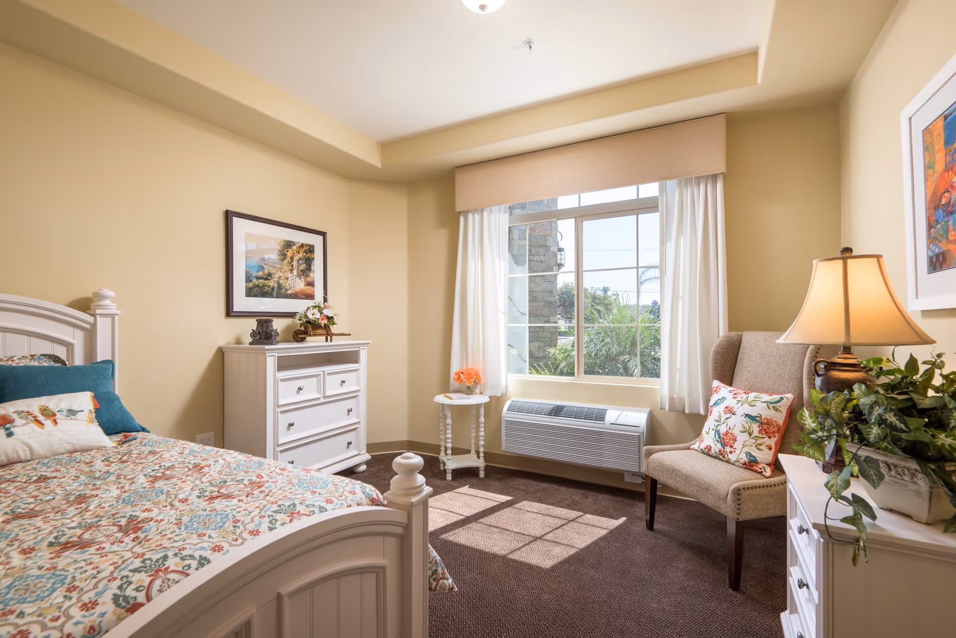 A bright and cozy bedroom with a white wooden bed featuring a colorful floral bedspread and pillows. There is a white dresser with decorative items and a framed picture above it. A window with white curtains lets in natural light, casting shadows on the carpeted floor. Next to the window is a small round table with a flower vase and a beige upholstered chair with a floral pillow. A lamp and a green plant sit on a white nightstand.