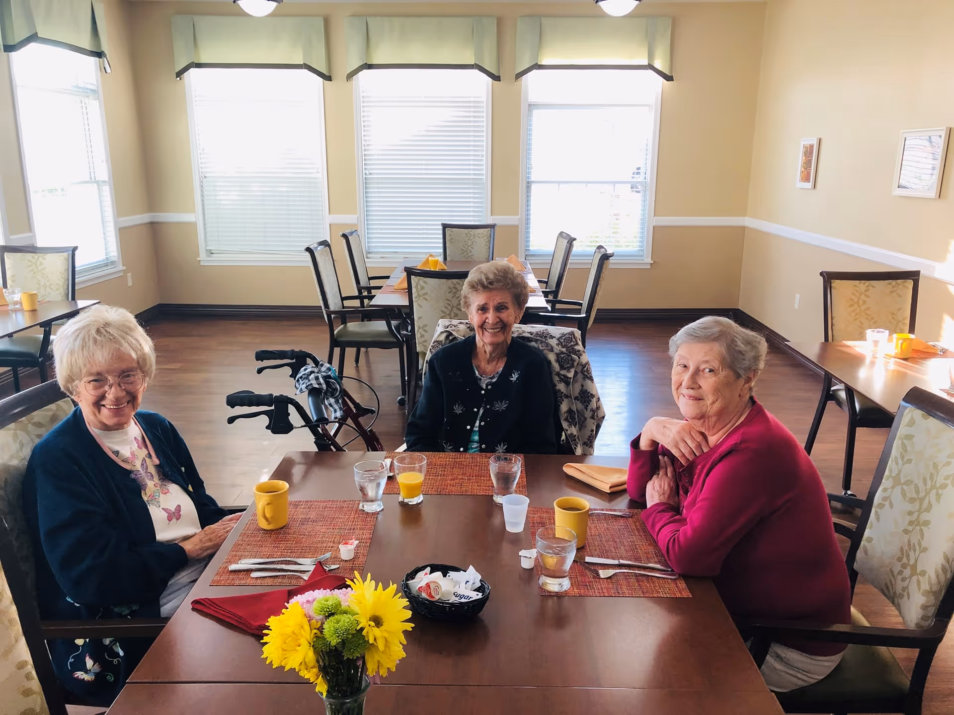 Three elderly women sitting around a dining table in a bright room with large windows. The table has placemats, cups, glasses, utensils, and a small vase with yellow flowers. One woman has a walker beside her chair. The room has light-colored walls and wooden flooring.