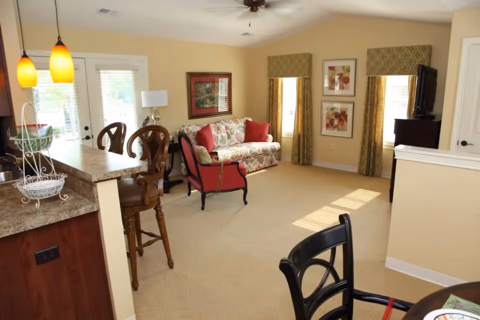A cozy living room area with a floral patterned sofa and red cushions, a red armchair, two framed pictures on the wall, and two windows with green patterned valances and curtains. The room has beige walls and carpet, a ceiling fan, and a TV on a dark wooden stand. To the left, there is a kitchen counter with two wooden bar stools and hanging pendant lights.