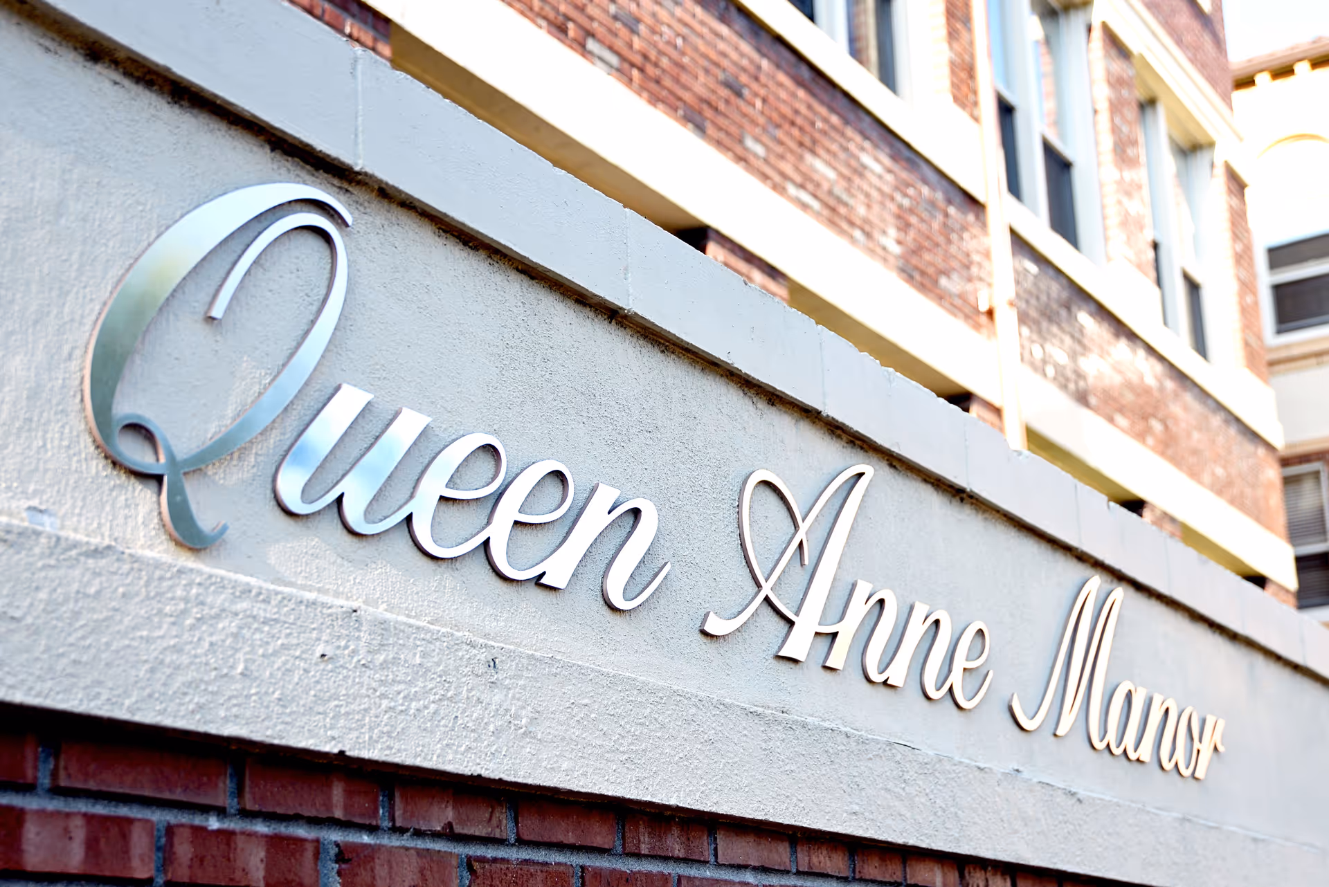 Close-up view of a building sign that reads 'Queen Anne Manor' mounted on a concrete wall with a brick building in the background.