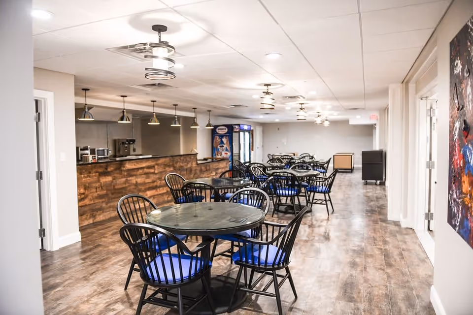 Large dining room with round glass-top tables, black chairs with blue cushions, a wooden service counter and pendant lighting.