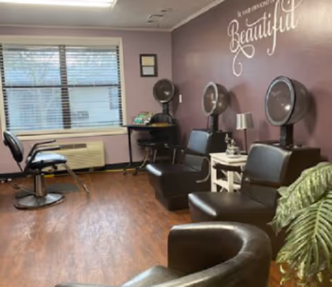 Interior of a salon area in Springfield Assisted Living with three black salon chairs, two hair dryers mounted on the wall, a small table with a lamp, and a window with blinds. The walls are painted purple with the phrase 'You are Beautiful' written in white cursive lettering.