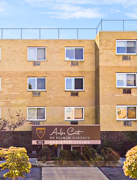 Exterior view of Amber Court of Pelham Gardens, a multi-story assisted living facility with beige brick walls and multiple windows. The entrance is marked by a sign displaying the facility's name and logo, surrounded by landscaped bushes and trees.