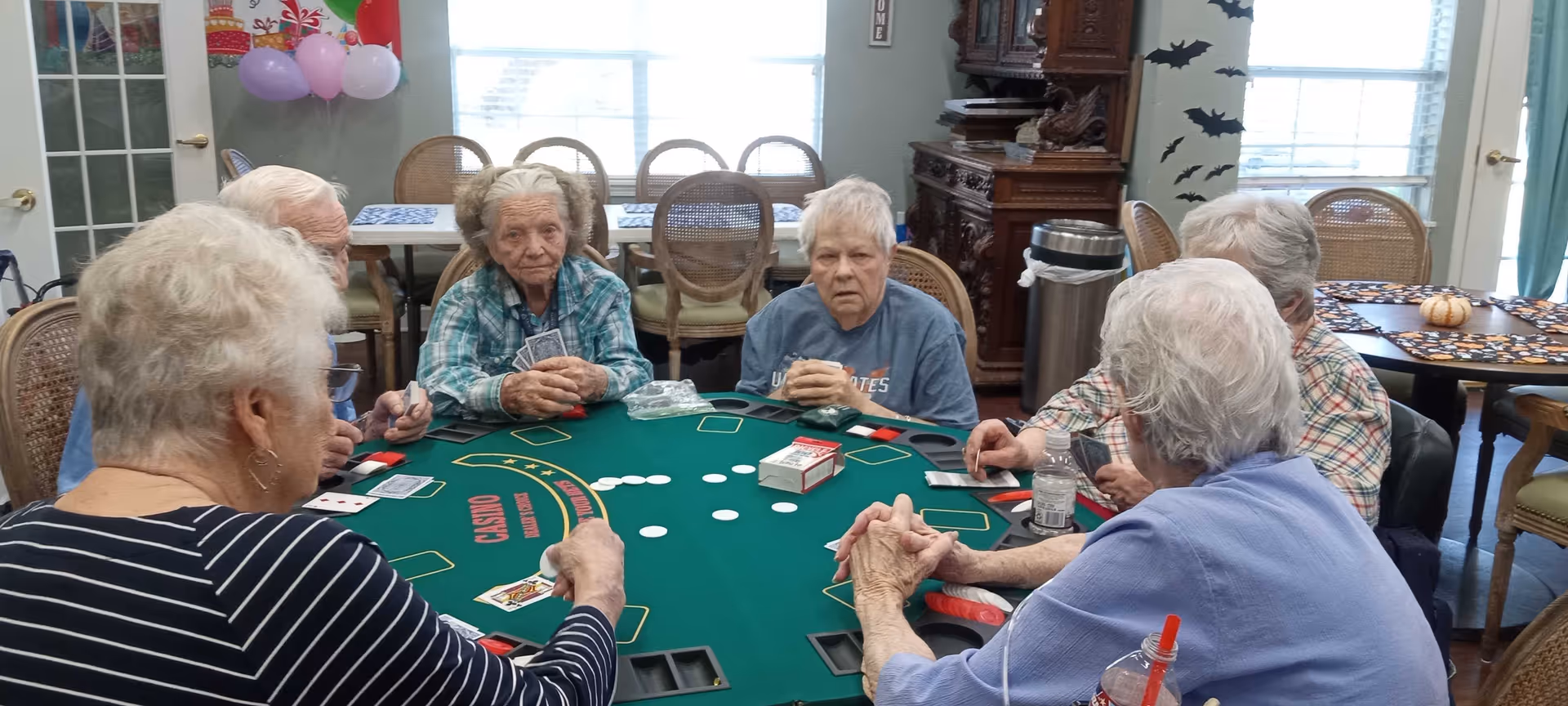 A group of elderly people sitting around a green casino-style card table playing cards in a well-lit room with windows, balloons, and Halloween decorations on the walls.