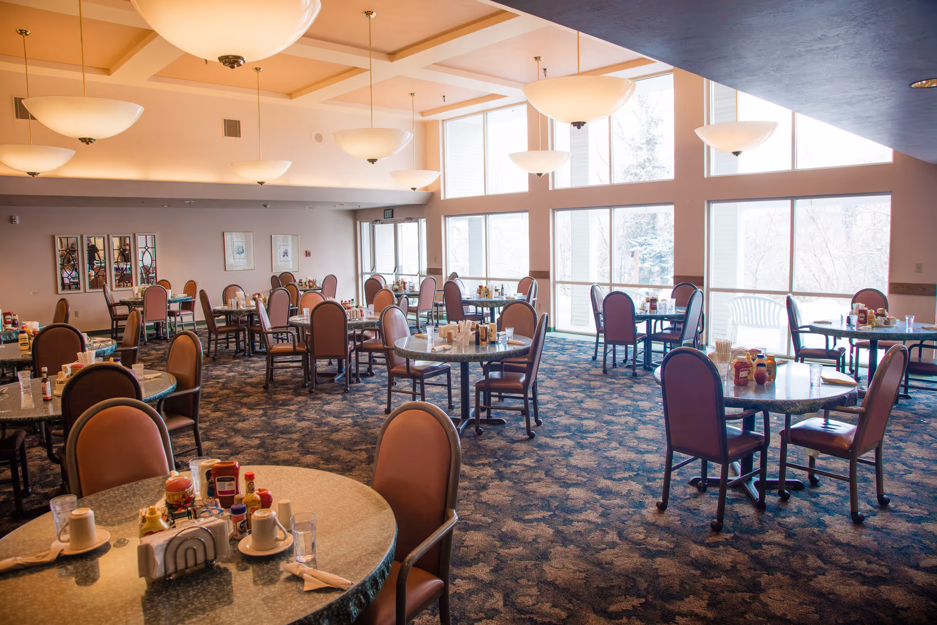 A spacious dining room with multiple round tables and chairs arranged neatly. Each table is set with condiments, napkins, cups, and glasses. Large windows on one side allow natural light to fill the room, and several ceiling lights hang from the ceiling. The room has a patterned carpet and light-colored walls with framed artwork.