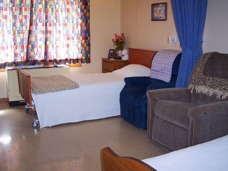 A small retirement facility bedroom with two beds, upholstered recliners, a bedside table with flowers and photos, and colorful curtains.