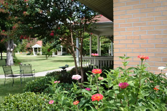 Flower bed of pink and red zinnias beside a brick building porch overlooking a grassy lawn with chairs and a distant gazebo.
