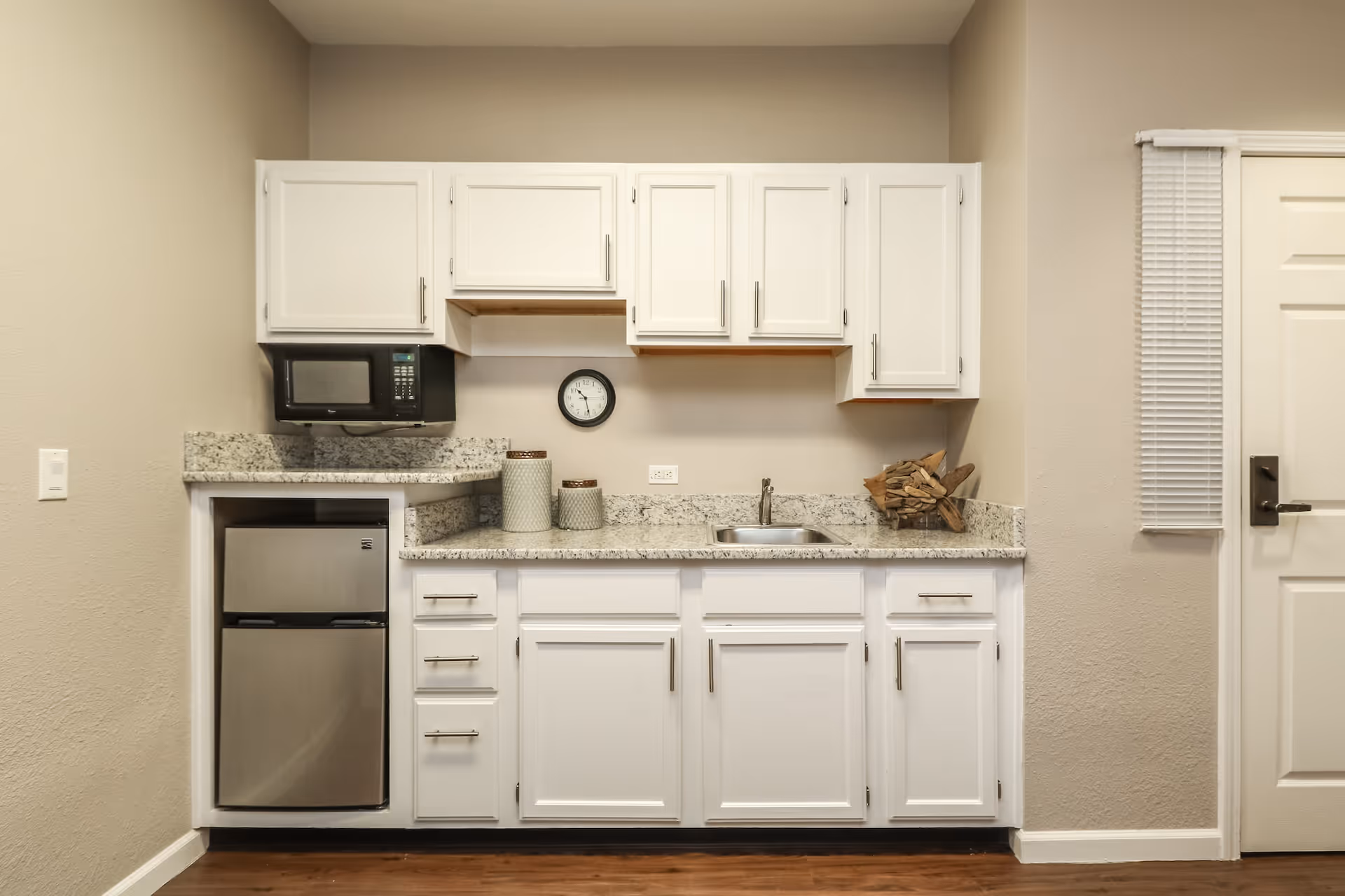 A small kitchen area with white cabinets, a granite countertop, a stainless steel mini refrigerator, a microwave, a small sink, and decorative containers. There is a clock on the wall above the countertop and a door with a window blind to the right.