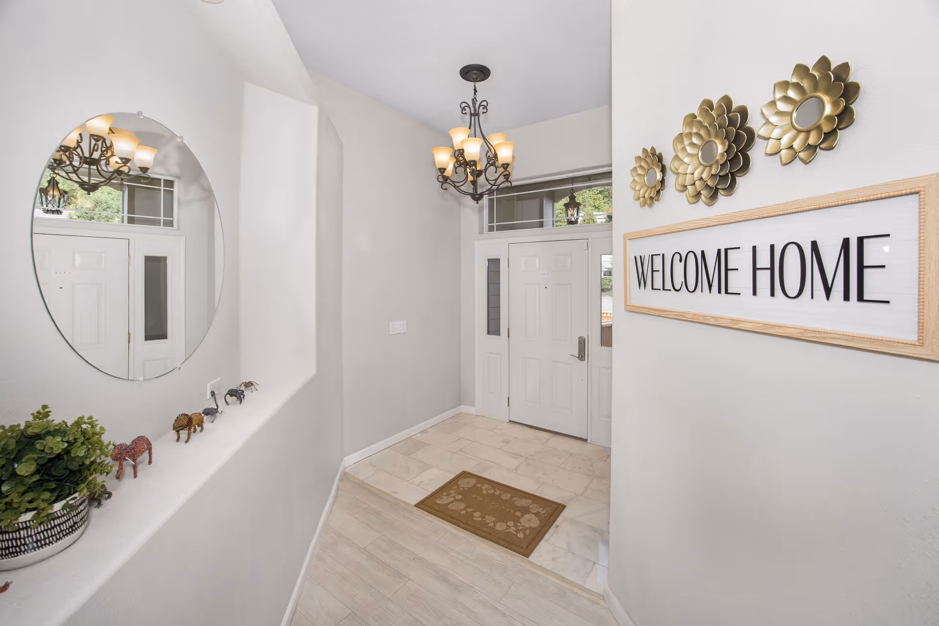 Bright entryway with a white front door, a 'WELCOME HOME' sign, round mirror, chandelier, and decorative accents.