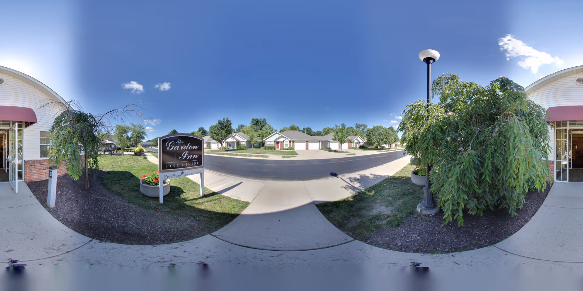 Outdoor view of a senior living facility entrance with a sign that reads 'The Garden Inn Fine Dining' and 'Five Star Residences'. The scene includes a paved sidewalk, green shrubs, a lamp post, and residential-style buildings in the background under a clear blue sky.