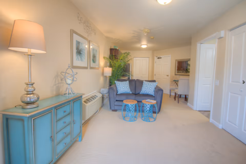 A cozy living room area in a senior living facility with a gray loveseat adorned with two patterned blue and white pillows. In front of the loveseat are two small round blue metal side tables with wooden tops. To the left, there is a turquoise sideboard with a silver lamp and decorative globe on top. The walls are beige with two framed abstract artworks and a decorative metal wall piece above the loveseat. A tall green plant is placed near the loveseat. The room has beige carpeting and white doors leading to other rooms, with a dining area visible in the background.