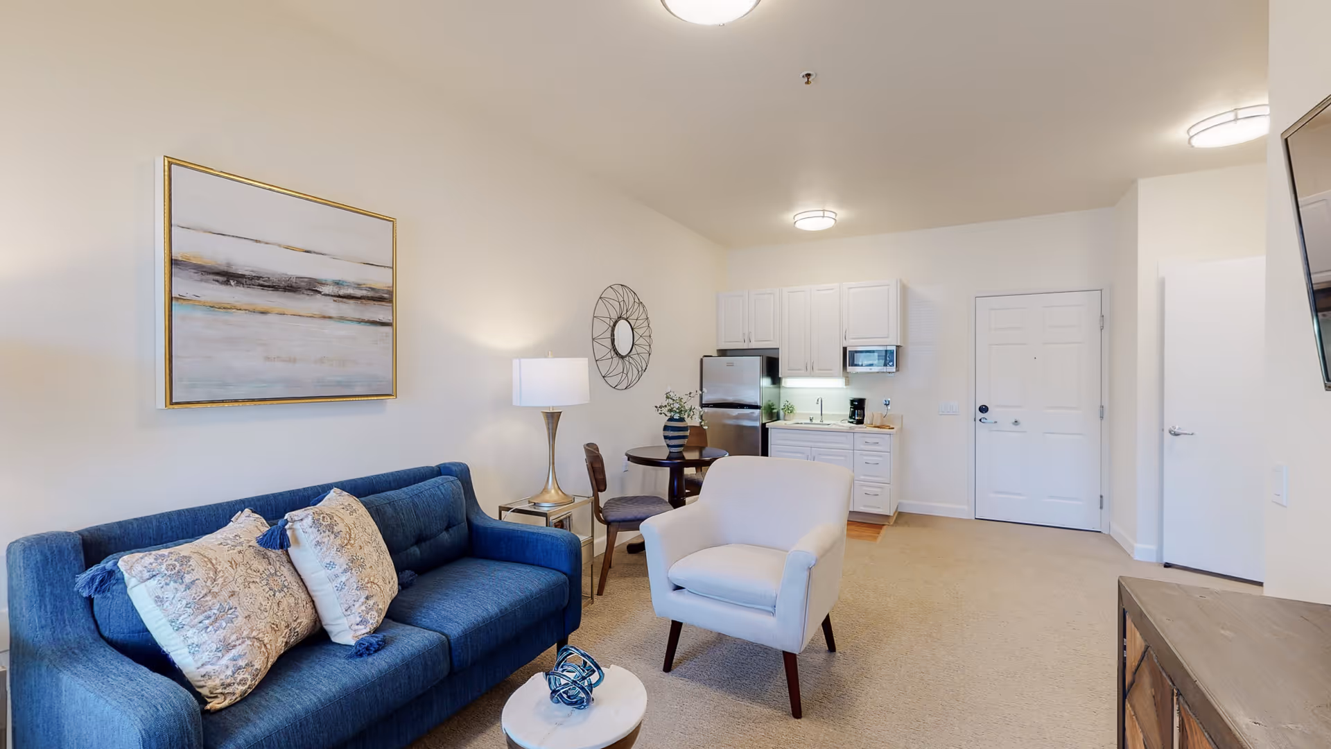 Open living area with a blue sofa, armchair, small dining table and a compact kitchenette with white cabinets.