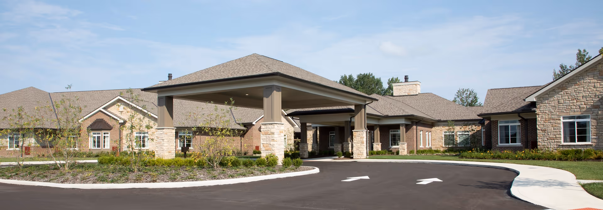 Front exterior view of The Ganzhorn Suites building with a covered entrance, brick and stone facade, landscaped greenery, and a paved driveway with directional arrows.