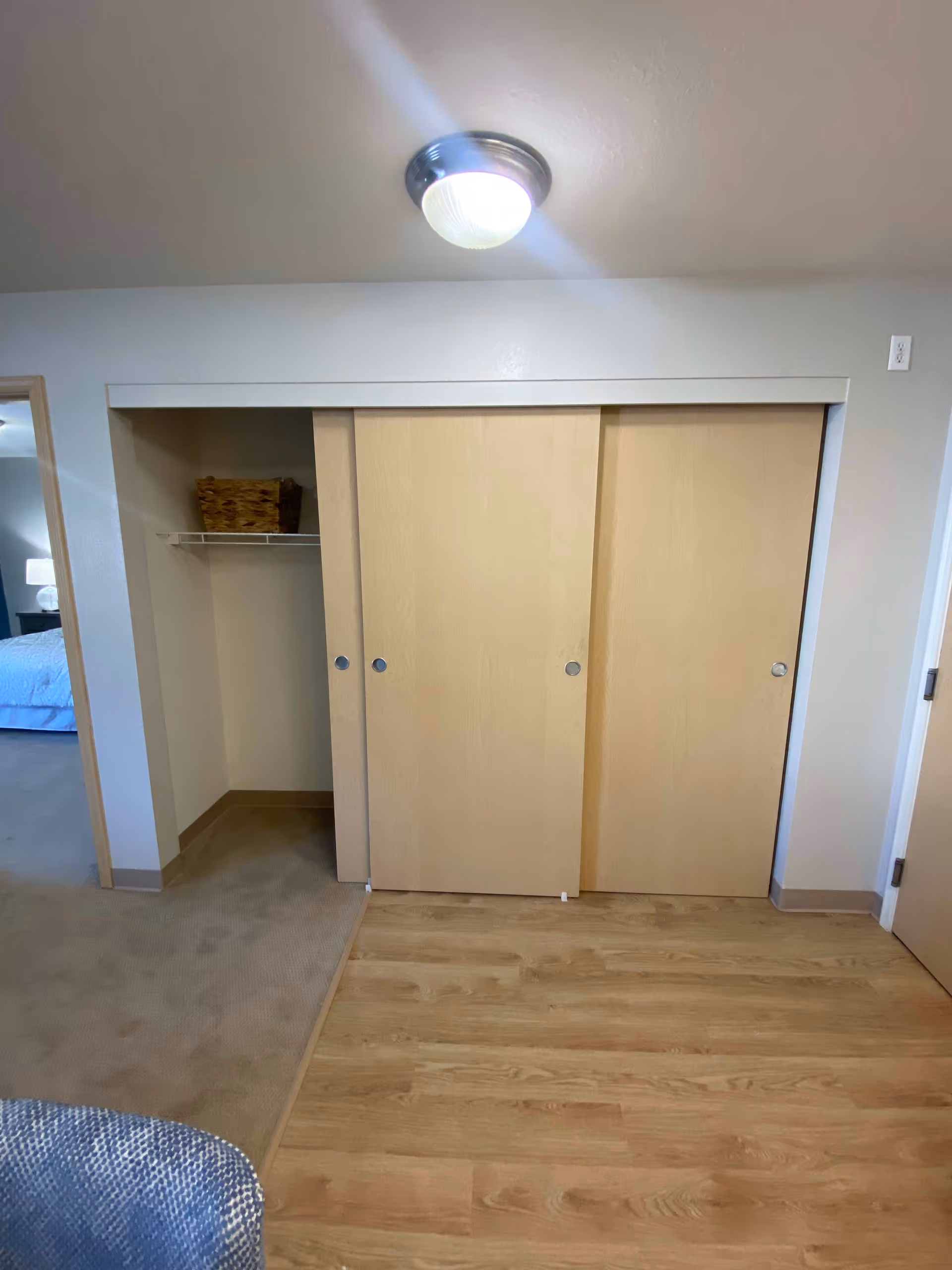 Interior view of a room with a partially open closet featuring sliding wooden doors. The closet has a shelf with a wicker basket. The floor is a combination of wood and carpet, and a ceiling light fixture is visible above. A glimpse of a bed and a lamp can be seen through an open doorway to the left.