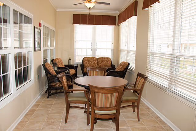 Sunlit common room with a round wooden table and chairs and wicker lounge seating by large windows.