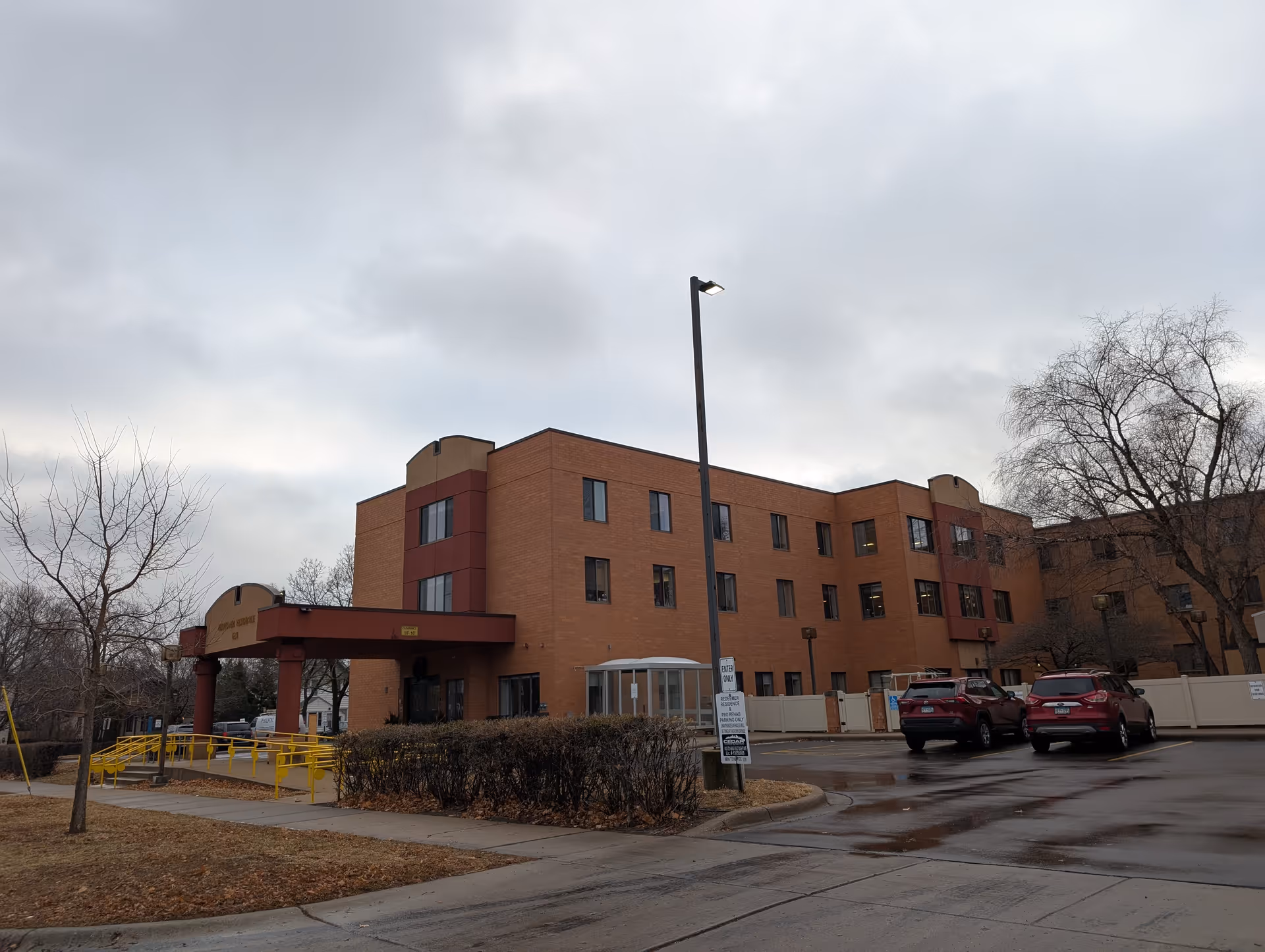 Exterior view of a three-story brick building with a covered entrance and a yellow handrail ramp. There are two red cars parked in the parking lot, leafless trees, and a cloudy sky overhead.
