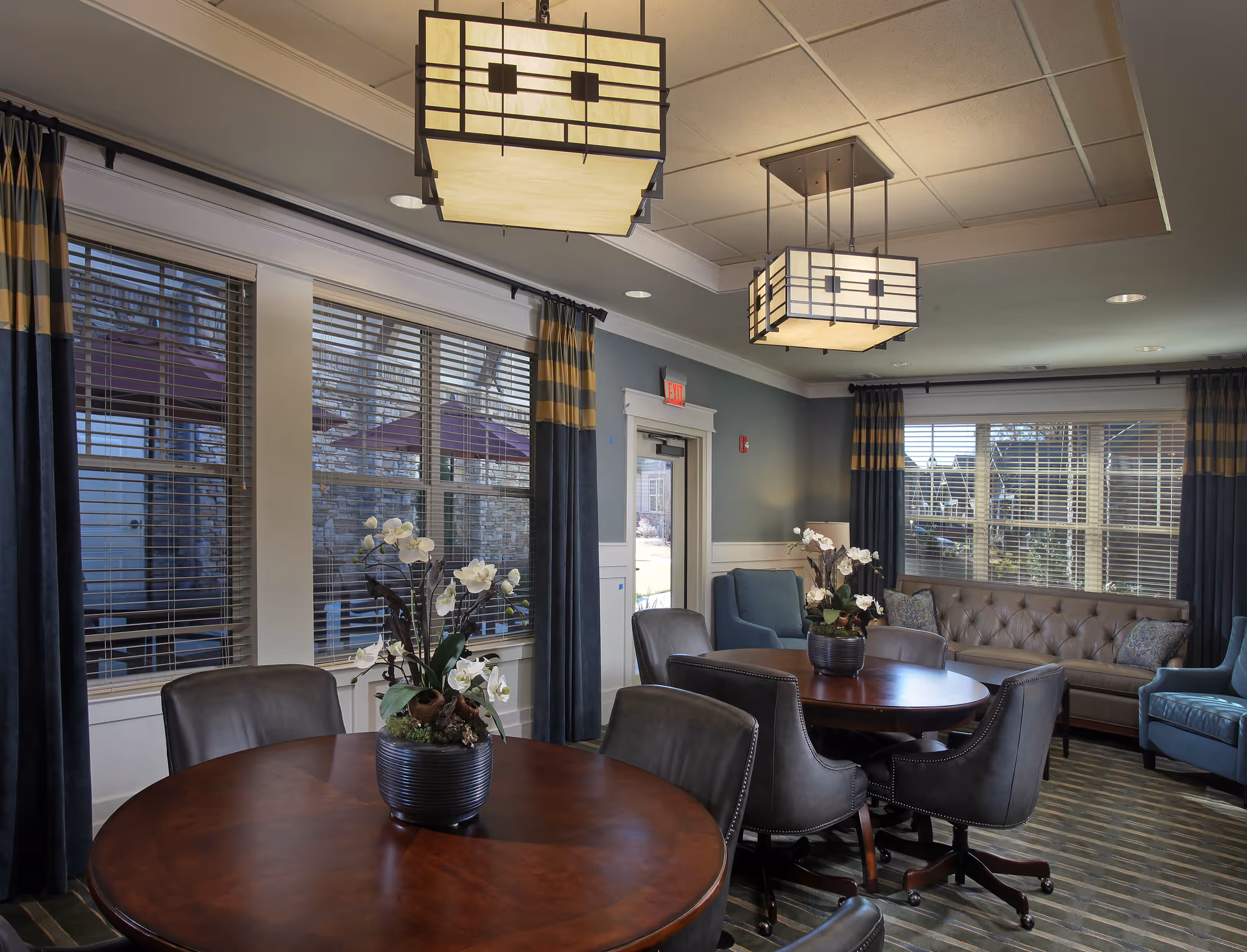 A cozy common area with round wooden tables surrounded by leather chairs, two large modern pendant lights hanging from the ceiling, large windows with striped curtains, and a tufted sofa along with armchairs near the windows.