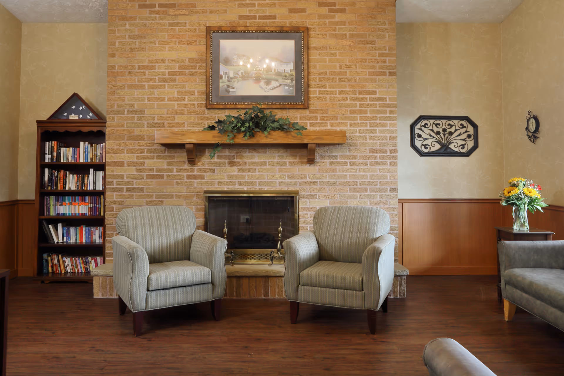 A cozy communal living room with a brick fireplace flanked by two striped armchairs, a bookshelf, wall decor, and a vase of flowers.