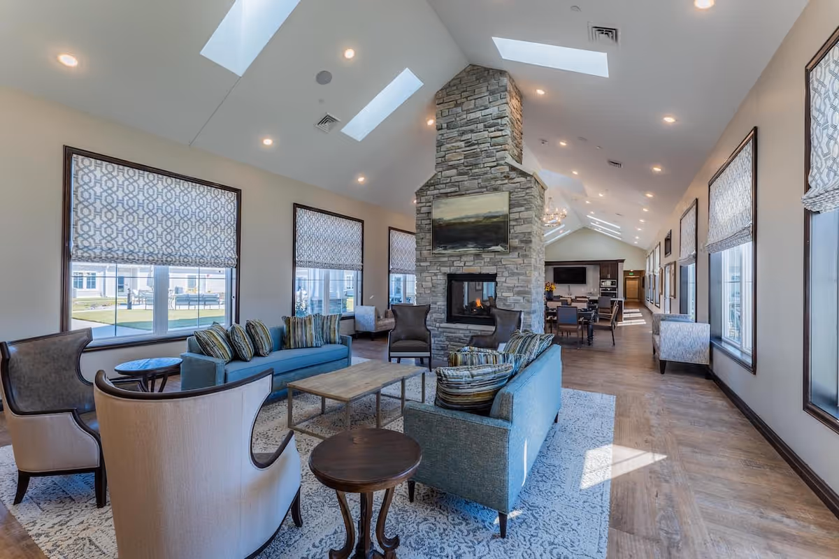 A spacious senior living facility common area with high vaulted ceilings and skylights. The room features a stone double-sided fireplace with a painting above it, surrounded by blue sofas, armchairs, and small wooden tables. Large windows with patterned blinds let in natural light, and a dining area with tables and chairs is visible in the background.