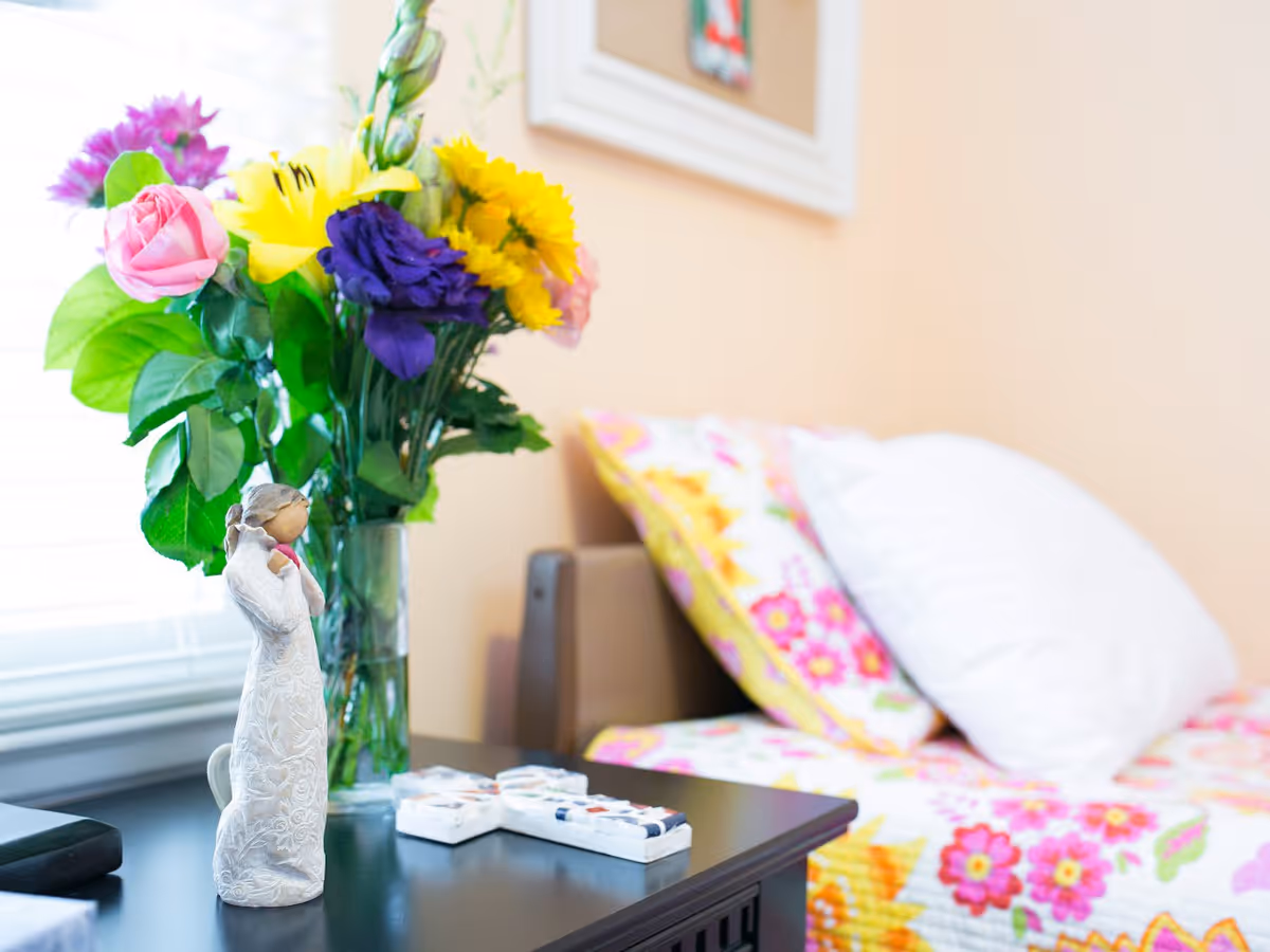 A close-up view of a bedside table with a decorative figurine, a vase of colorful flowers including yellow, purple, and pink blooms, and a bed with floral-patterned bedding and pillows in the background.