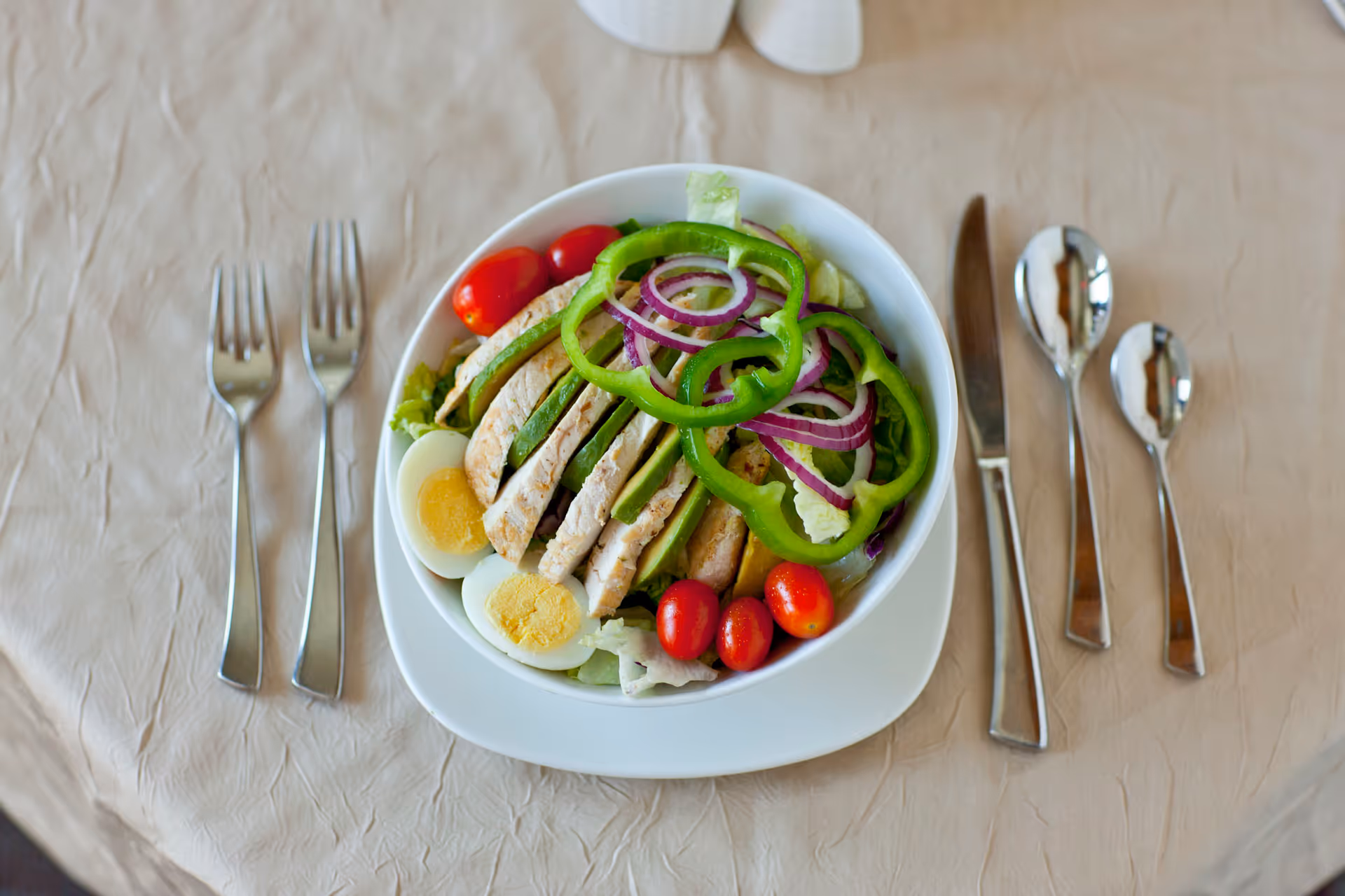 A bowl of salad containing sliced grilled chicken, green bell pepper rings, red onion rings, cherry tomatoes, lettuce, and hard-boiled egg halves, placed on a white plate on a beige tablecloth. The table is set with two forks on the left and a knife, a large spoon, and a small spoon on the right.