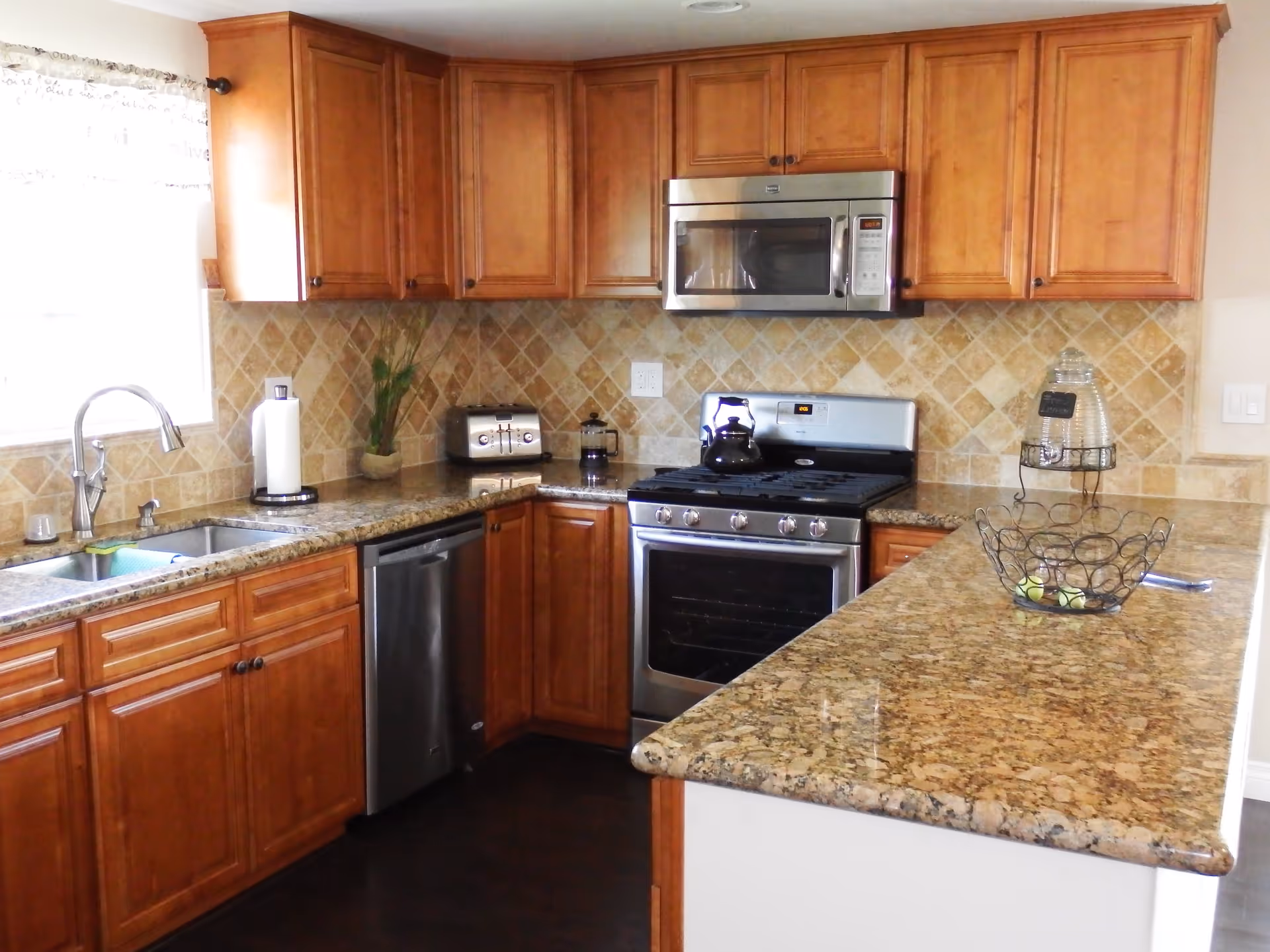 A kitchen with wooden cabinets, granite countertops, a stainless steel sink, dishwasher, stove with oven, and microwave. There is a paper towel holder, toaster, French press, and a decorative plant on the countertop. A wire fruit basket and a glass water dispenser are on the island countertop.
