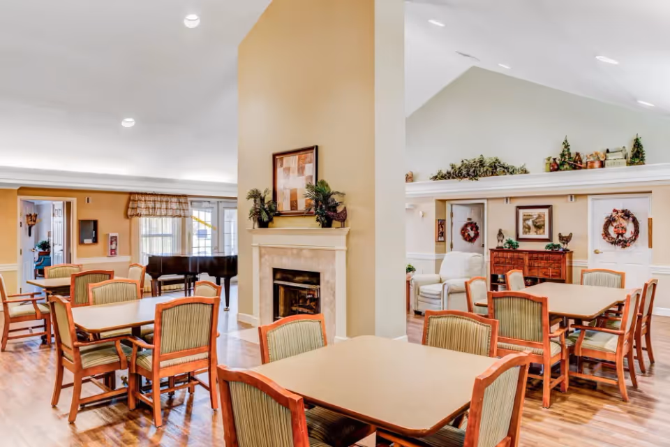 A spacious common area with multiple tables and chairs arranged for dining or socializing. The room features a central fireplace with decorative plants and artwork above it. There is a grand piano near the window with curtains, and the walls are adorned with wreaths and framed pictures. The flooring is wooden, and the ceiling is vaulted with recessed lighting.