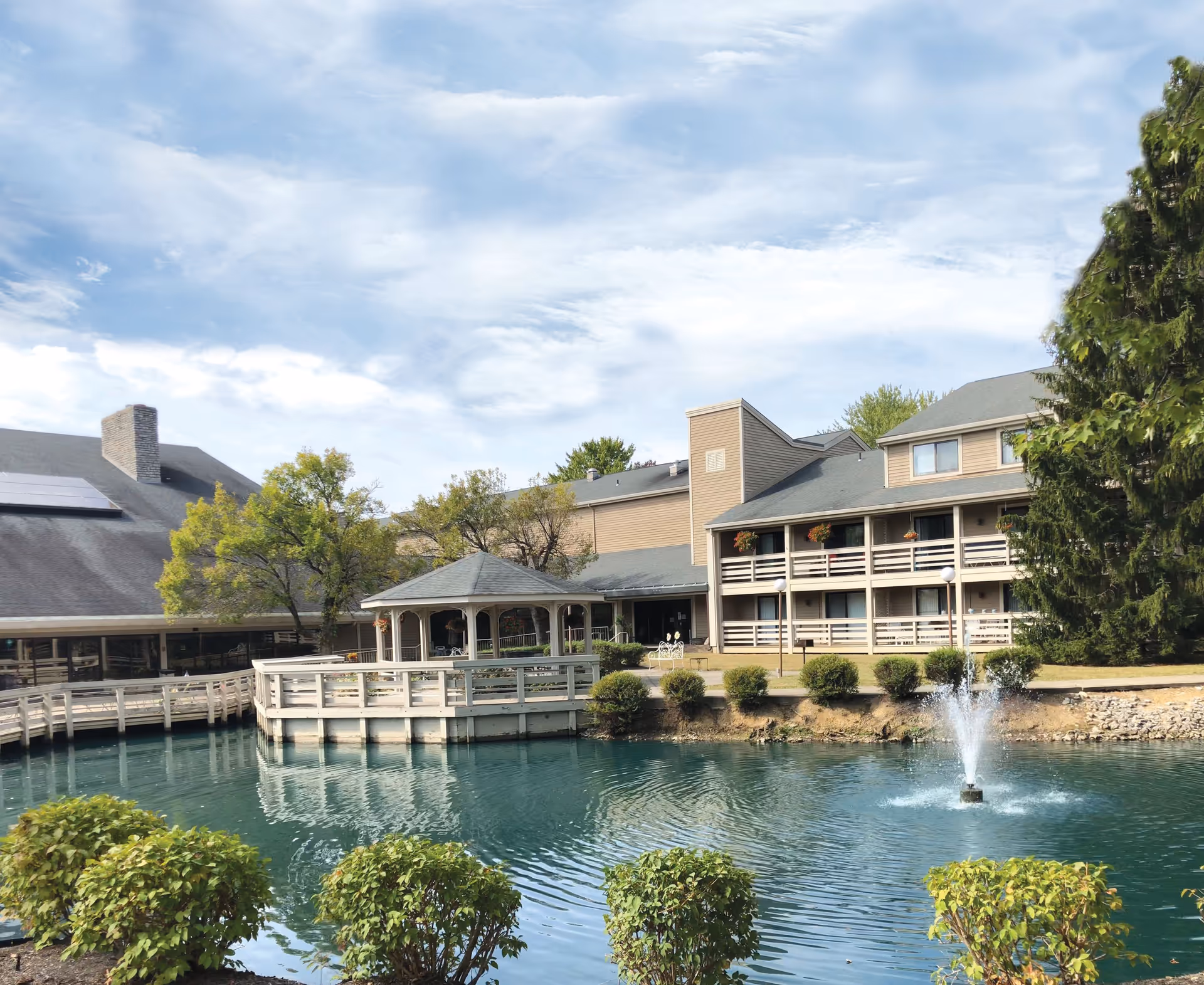 View of The Lodge Retirement Community showing a large pond with a fountain in the foreground, surrounded by bushes. Behind the pond is a wooden gazebo and a multi-story building with balconies and hanging flower pots. Trees and a partly cloudy sky are visible in the background.