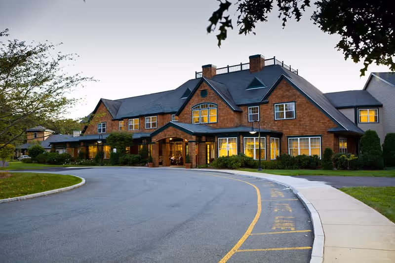 Exterior view of a large two-story senior living facility building with brown shingle siding and multiple windows lit from inside during dusk. The building has a covered entrance with a driveway and landscaped greenery around it.