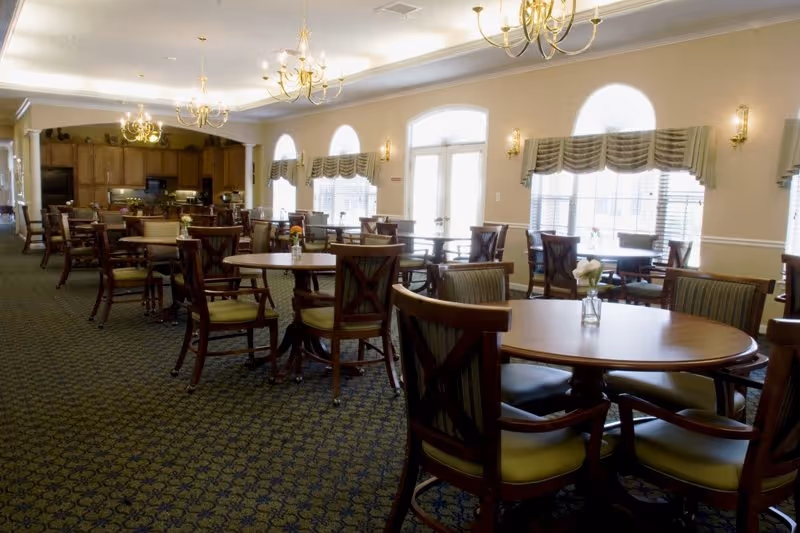 A spacious dining room with multiple round wooden tables and cushioned chairs arranged neatly. The room features patterned carpet flooring, large windows with valances allowing natural light, and chandeliers hanging from the ceiling. In the background, there is a kitchen area with wooden cabinetry and appliances.