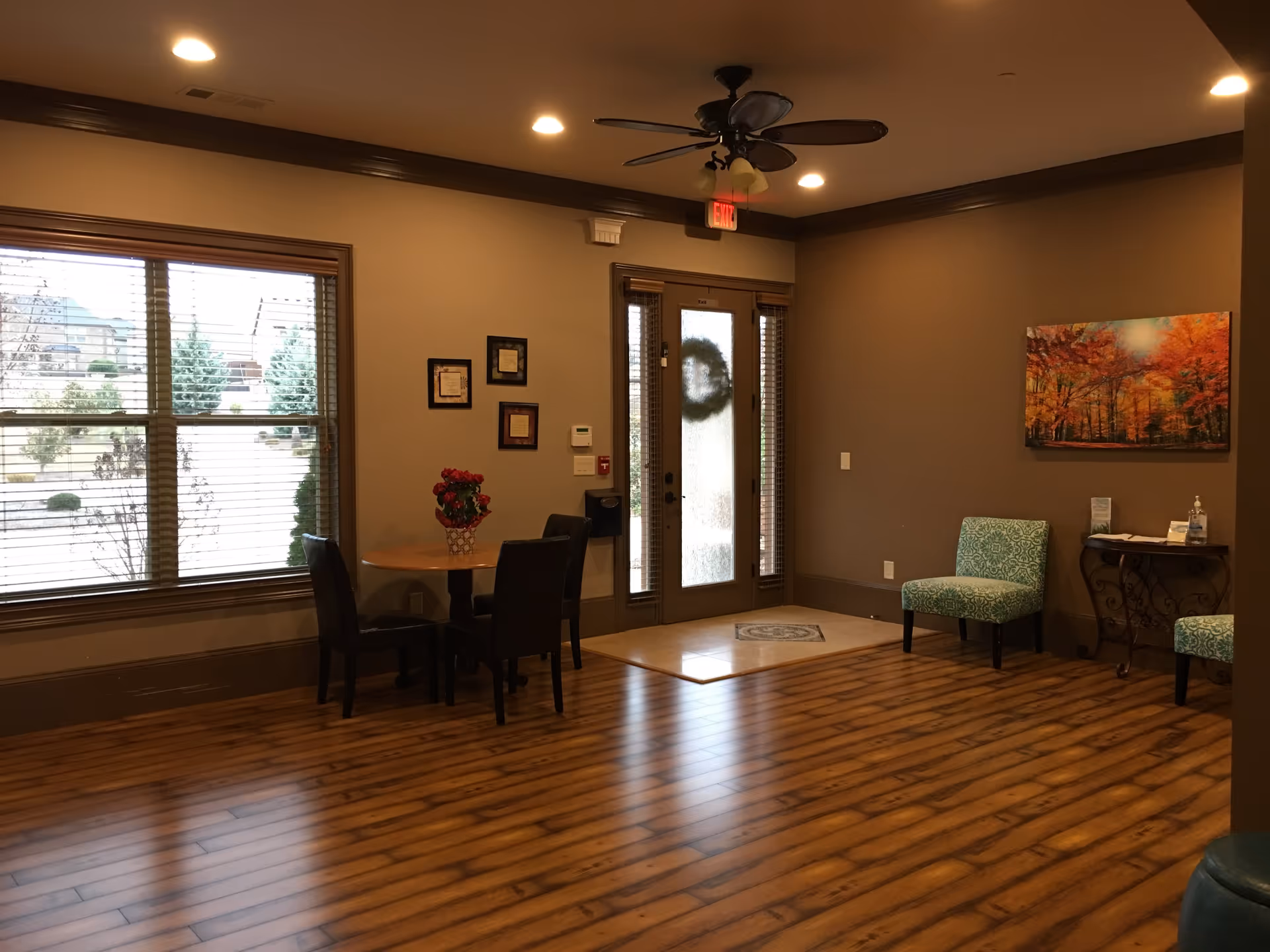 Interior view of a senior living facility's common area with wooden flooring, a ceiling fan with lights, a glass door with a wreath, a window with blinds, a small round table with two chairs and a flower vase, two patterned chairs, a small decorative table with hand sanitizer, and a colorful autumn forest painting on the wall.
