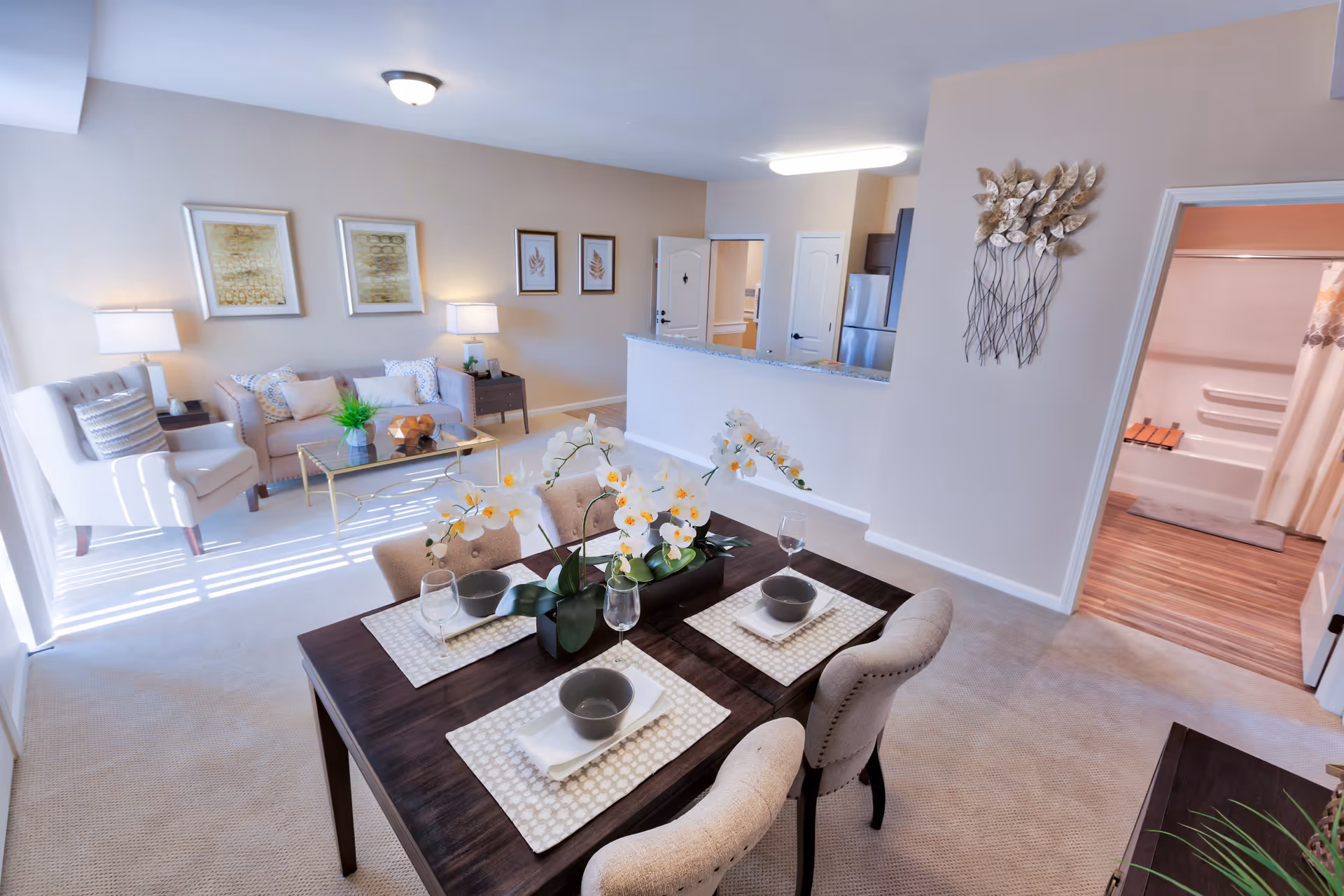 A bright and spacious senior living apartment interior featuring a dining table set for four with white orchids as centerpiece, a cozy living area with a sofa, armchair, coffee table, and lamps, and a kitchen with stainless steel appliances. A bathroom with a shower and curtain is visible through an open door.