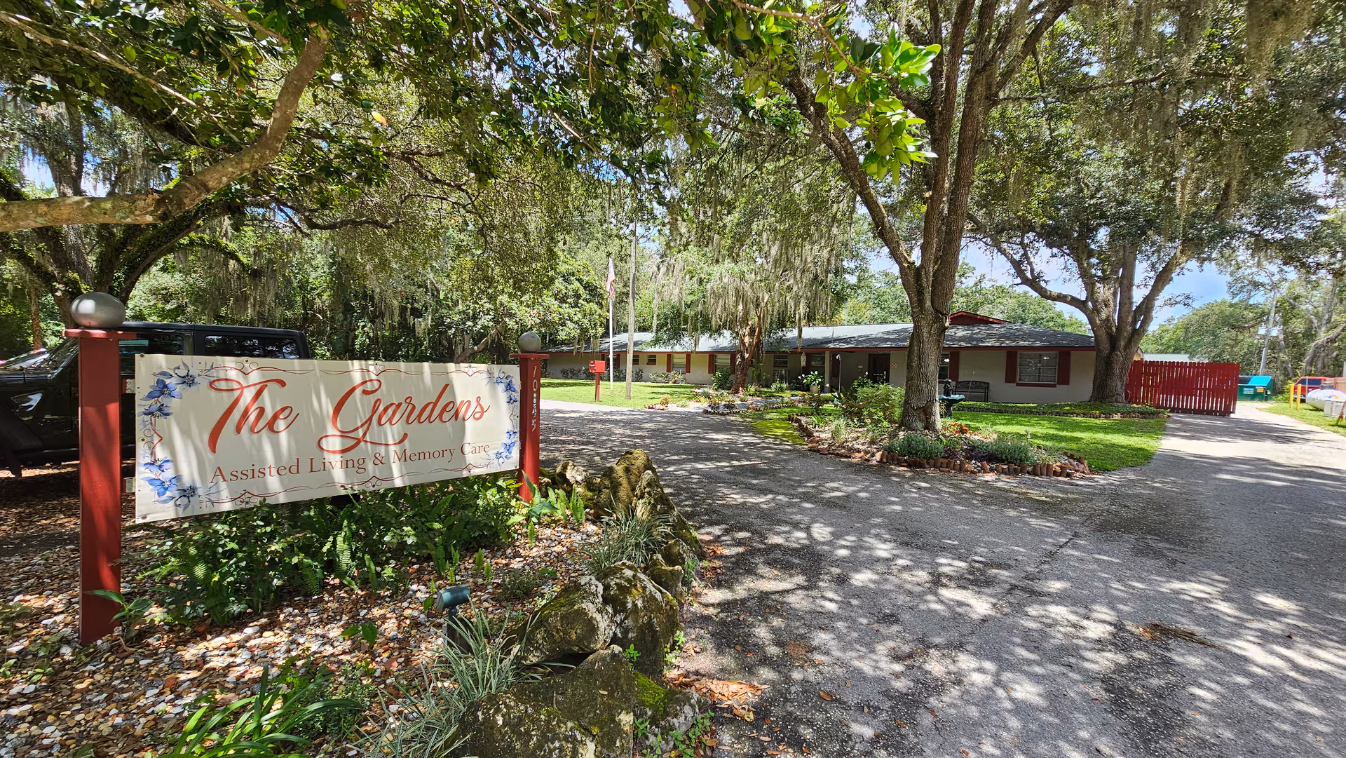 Entrance to The Gardens Assisted Living & Memory Care facility with a large sign displaying the facility name. The area is surrounded by trees and greenery, with a driveway leading to a single-story building in the background under a partly cloudy sky.