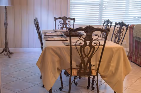 Dining area with a rectangular table covered with a beige tablecloth and set with placemats. The table is surrounded by ornate metal chairs with intricate back designs. The room has tiled flooring, a floor lamp, and large windows with white blinds letting in natural light.